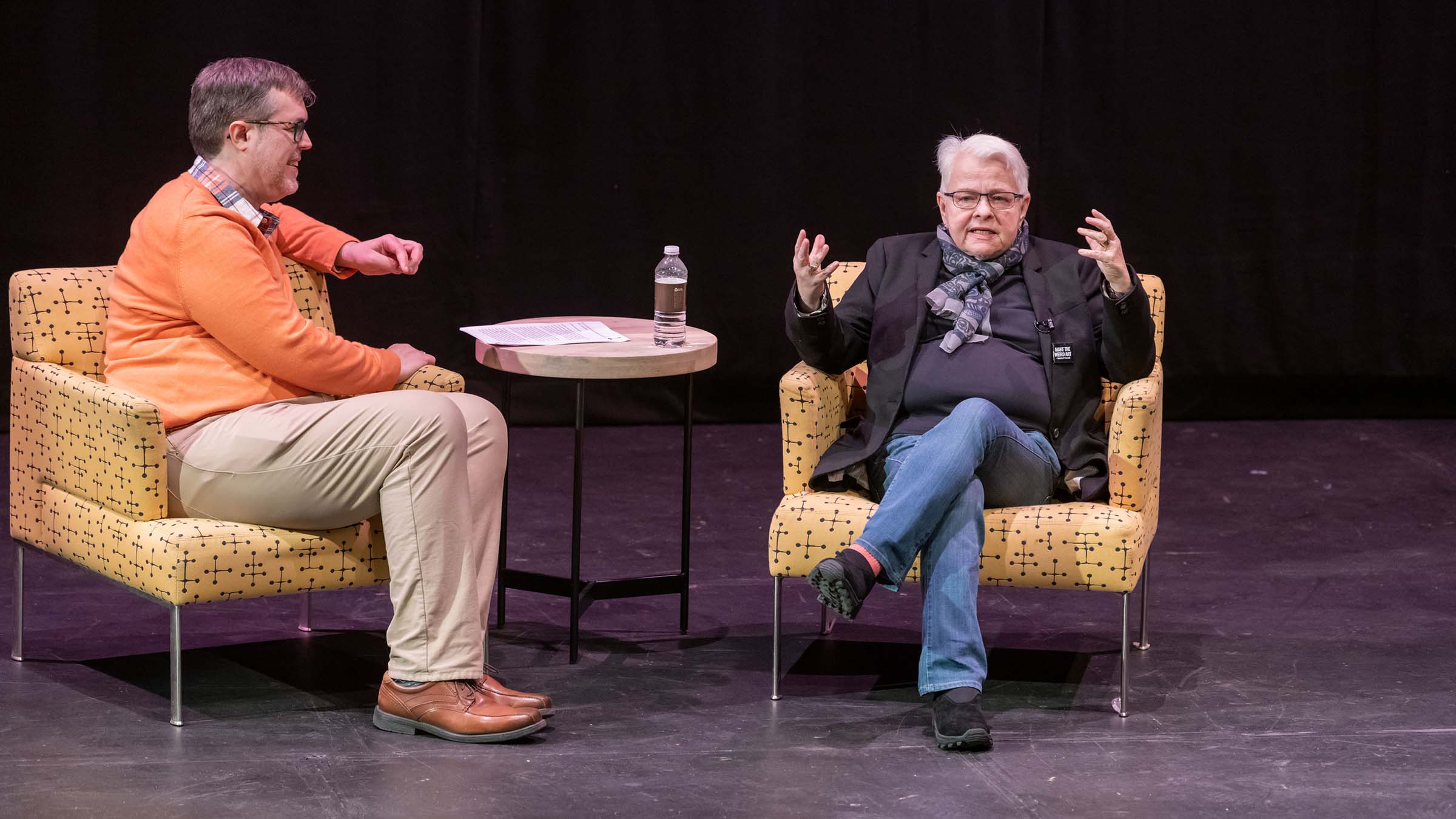 Assistant Professor of Drama Peter Gil-Sheridan wearing an orange sweater and tan pants and Pulitzer Prize-winning playwright Paula Vogel wearing a black shirt and jacket and jeans both sitting in a chair in the Martel Theater