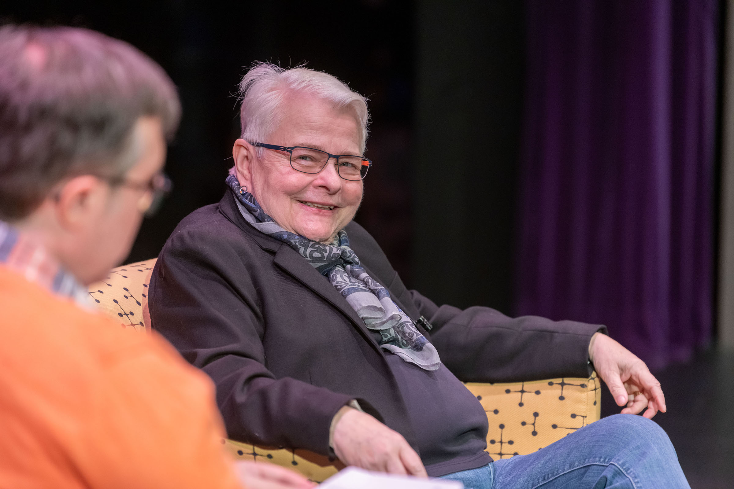 Pulitzer Prize-winning playwright Paula Vogel wearing a black shirt and jacket and jeans sitting in a chair in the Martel Theater