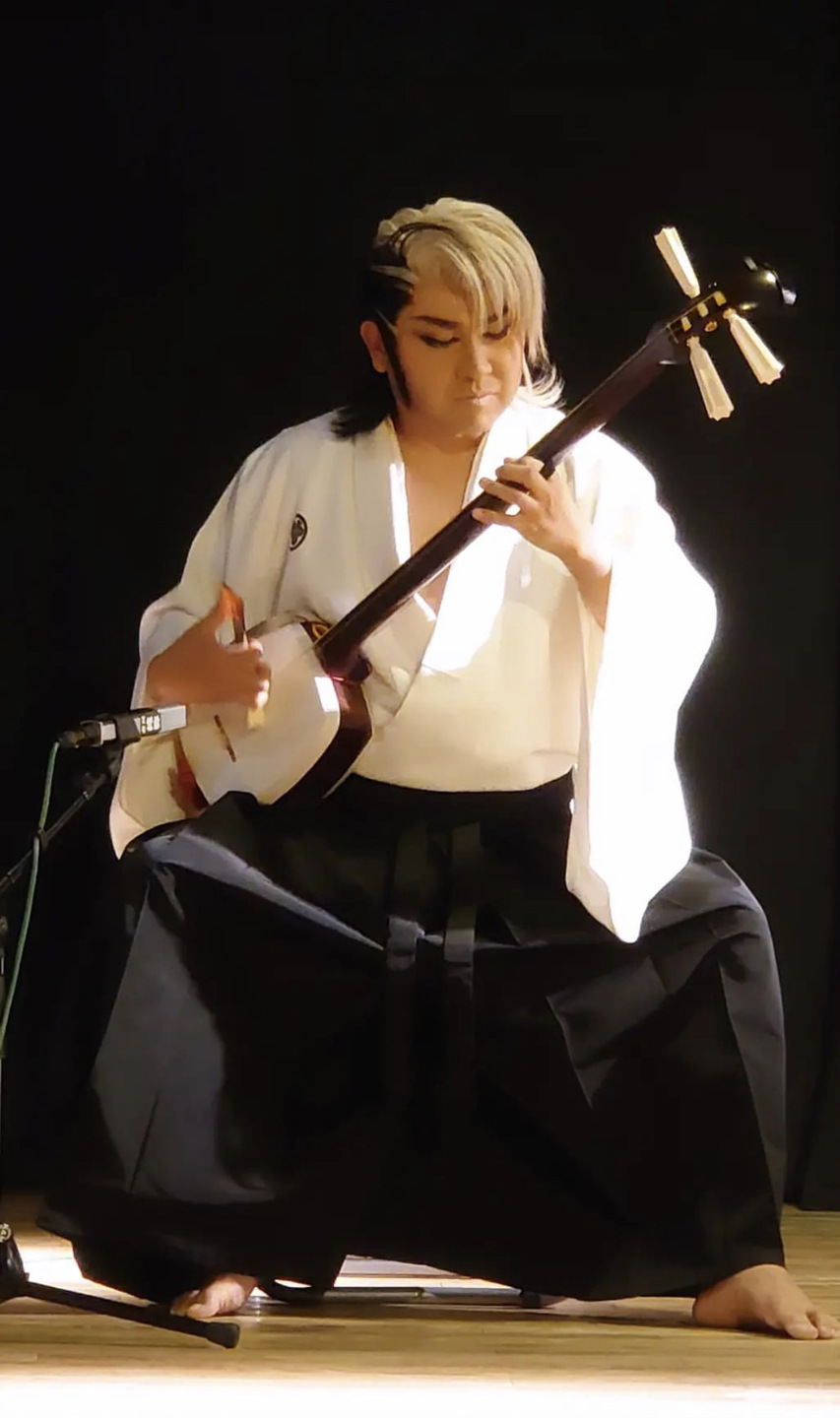 A seated, barefoot performer wearing robes strums a stringed instrument.