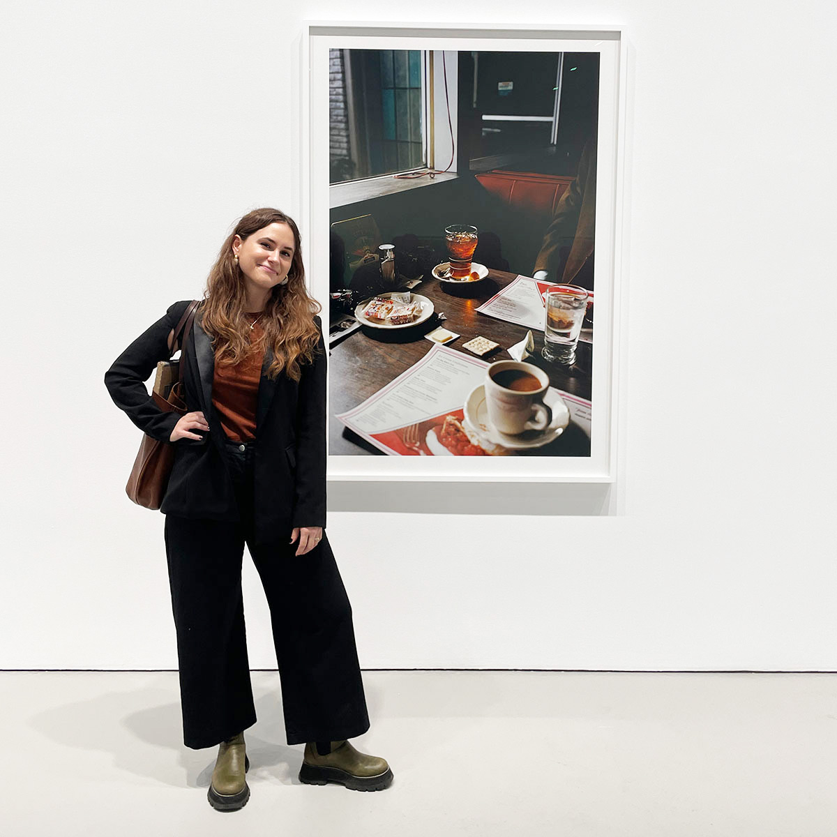 Nicola Turner wearing a black jacket and black pants standing by a white wall with a framed photo of a diner table with menu's, coffee, drinks and crackers.