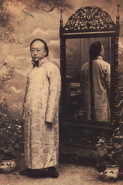 A person wearing a long silk robe with a flower pattern stands with his back to a mirror, which reflects his very long hair.