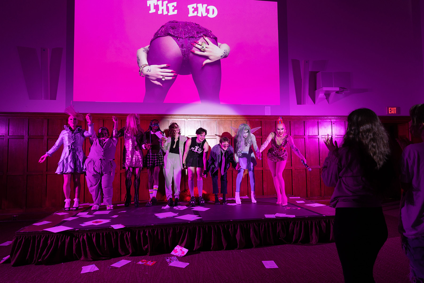 Performers on a dark pink stage bent over highlighting their backsides.