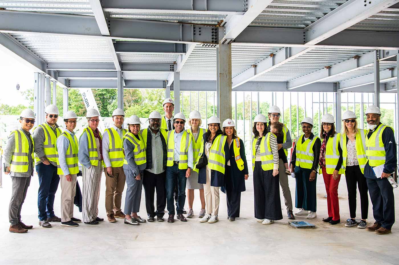 A group of nearly 20 people stand in an unfinished building that is being constructed, smiling at the camera, wearing fluorescent green vests and helmets.