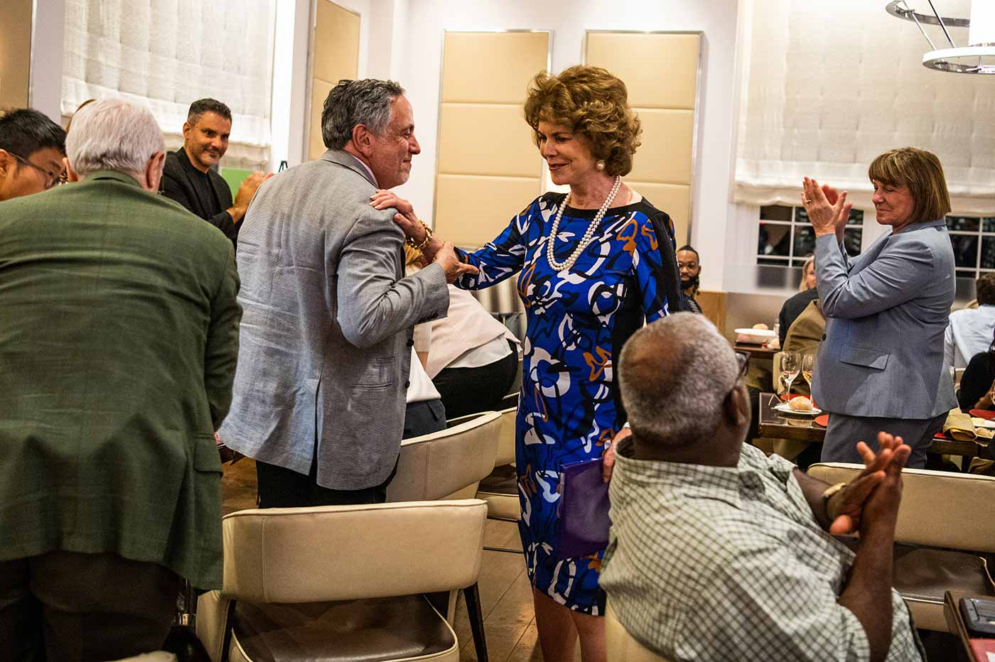 A group of people in formal attire applaud a person with mid-length brown hair, large pearl earrings, a pearl necklace, and a blue patterned dress. This is in a room, during a formal occasion.