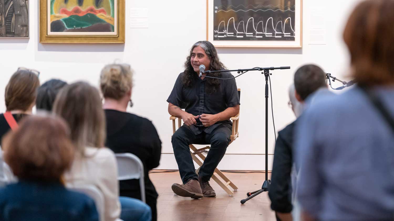 Person seated behind a microphone in an art gallery facing the camera and speaking to a seated audience