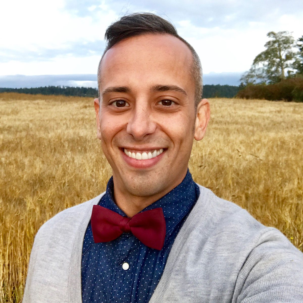 Justin Schantz wearing a blue printed shirt, gray sweater, red bowtie standing in a field.