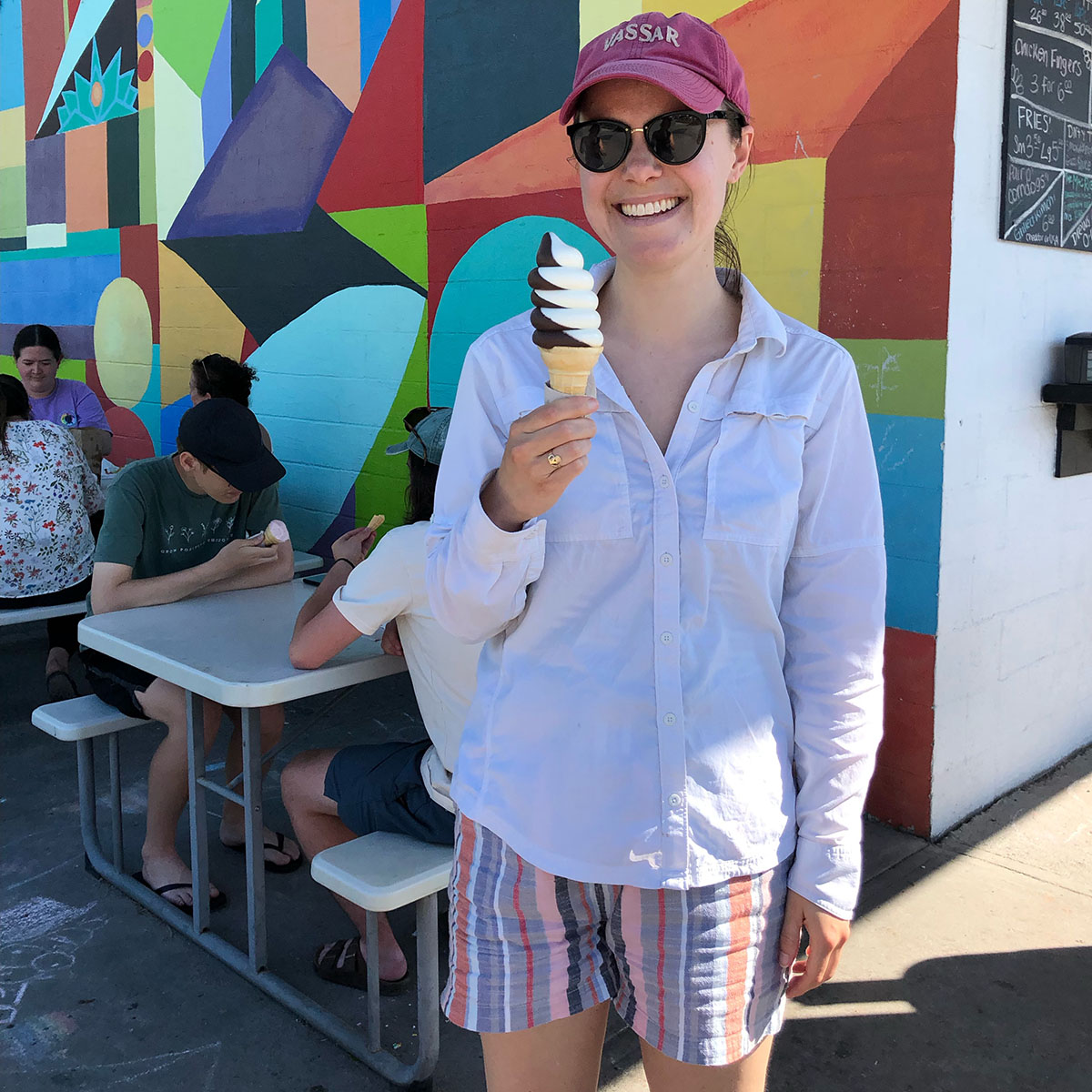 Sarah Scott wearing a Vassar hat, sunglasses, white shirt and striped colored shorts standing with an ice cream cone in front of a mural with people sitting on picnic tables.