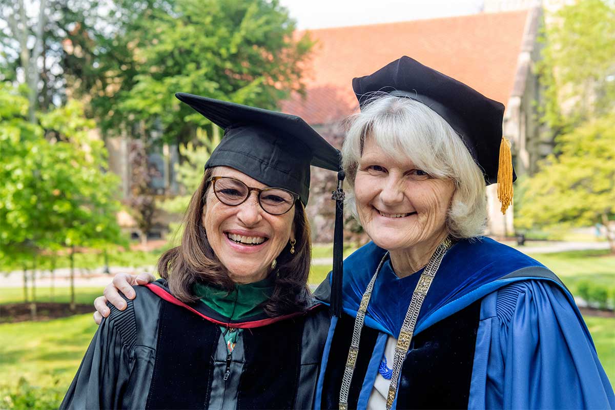 Two people in formal academic garb—robes and hats—smile at the camera. The person on the left has long brown hair, glasses, and a black robe; the person on the right has long gray hair and a blue robe.