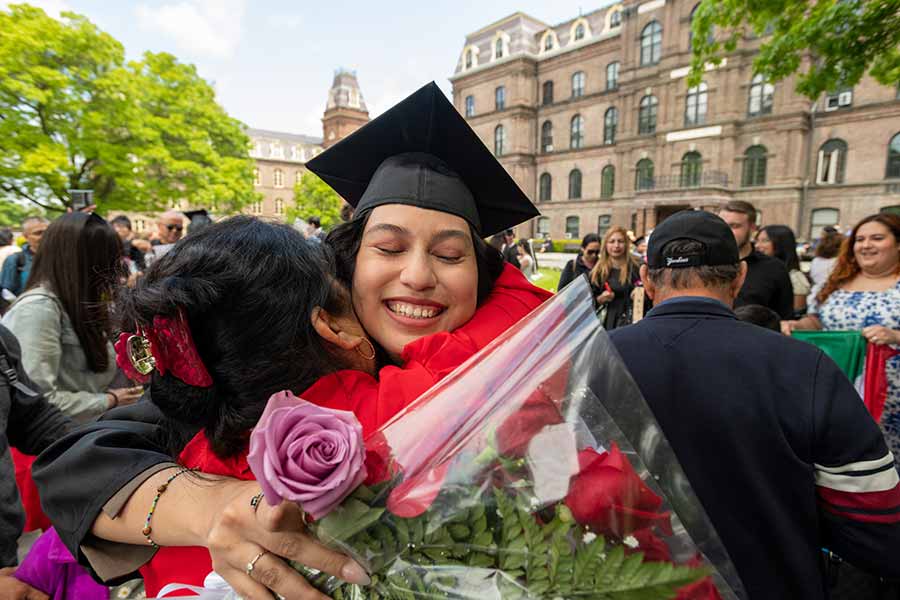 A student holding flowers and wearing a graduation robe hugs their mother in front of a crowd in front of Main Building, a large, multi-story, brick building.
