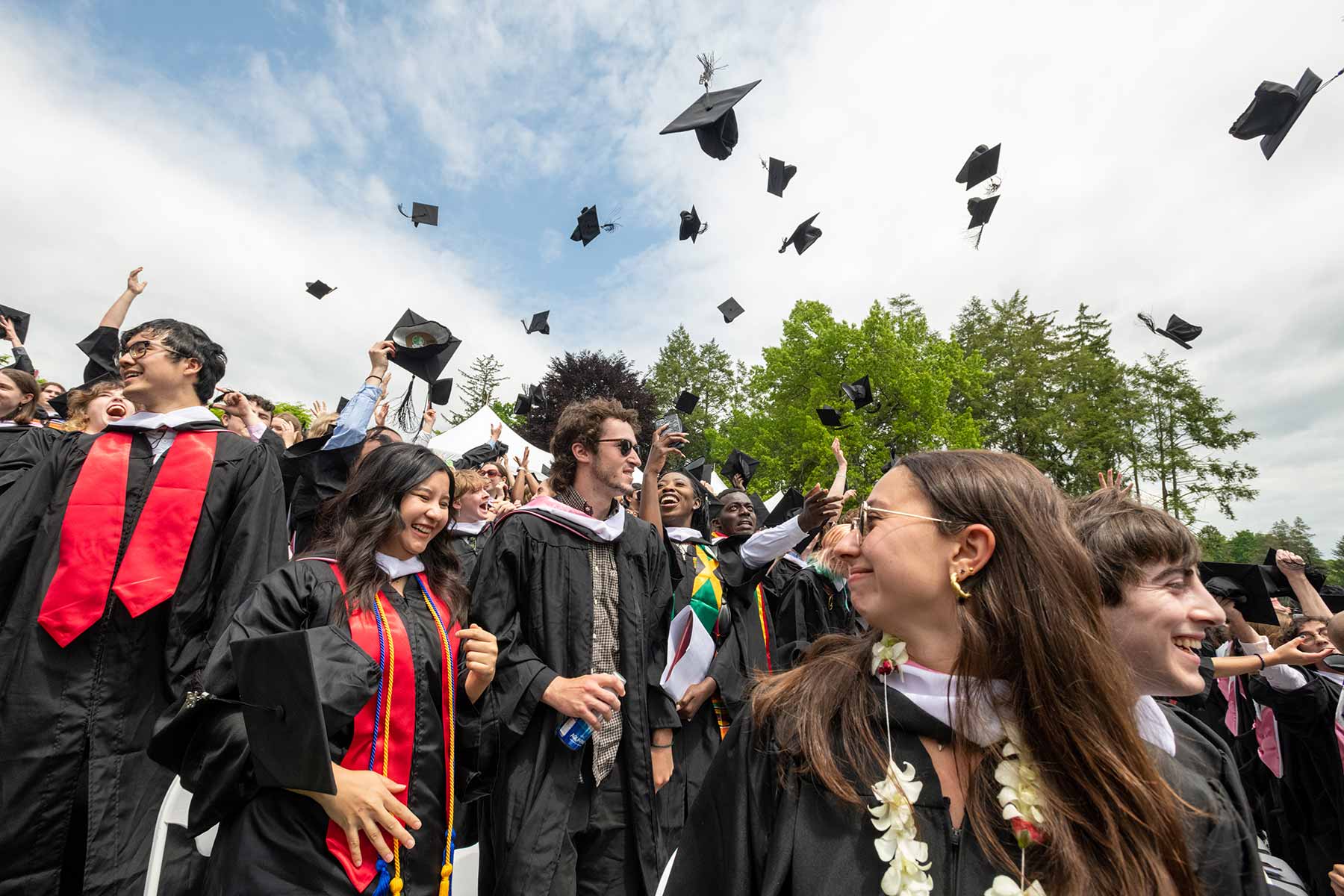 A group of people wearing dark graduation robes stand outside on a sunny day, tossing their hats into the air.