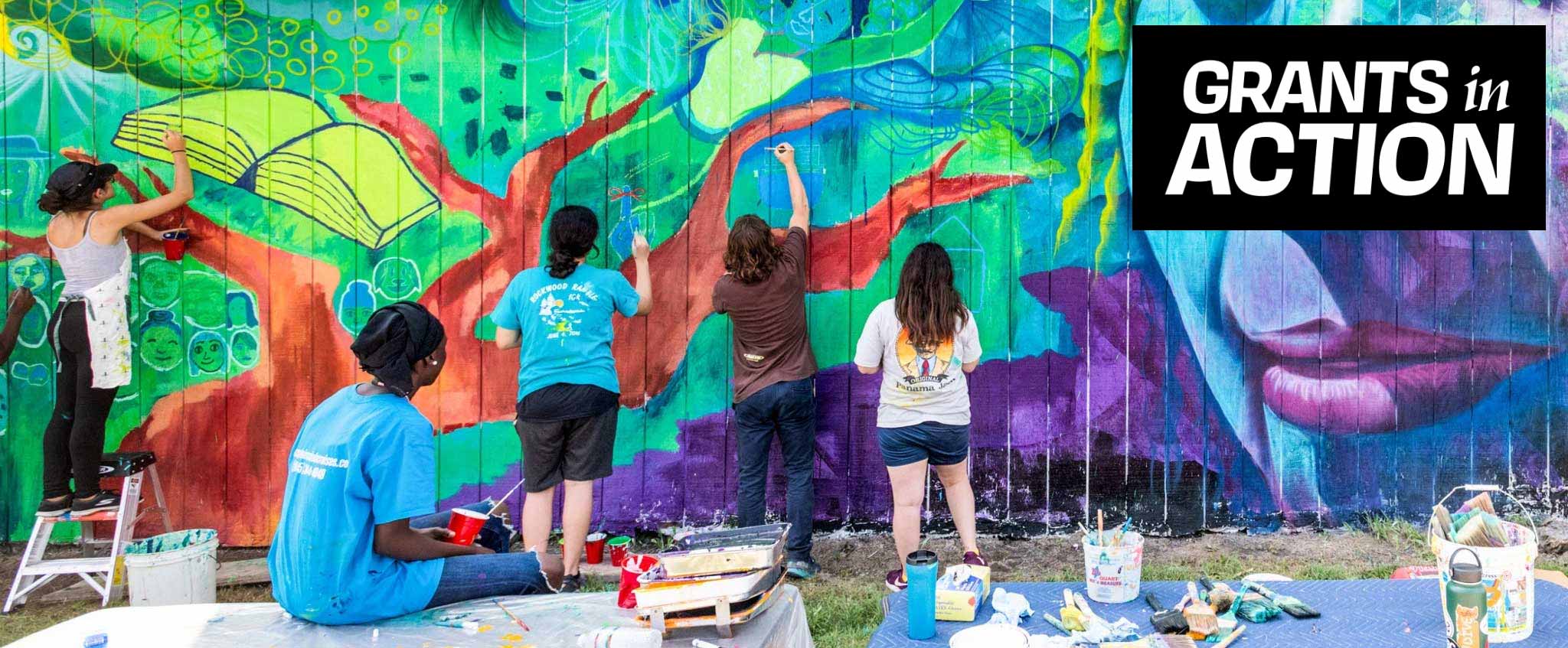 A group of people paint a brightly colored mural on the side of a building, backs facing the camera