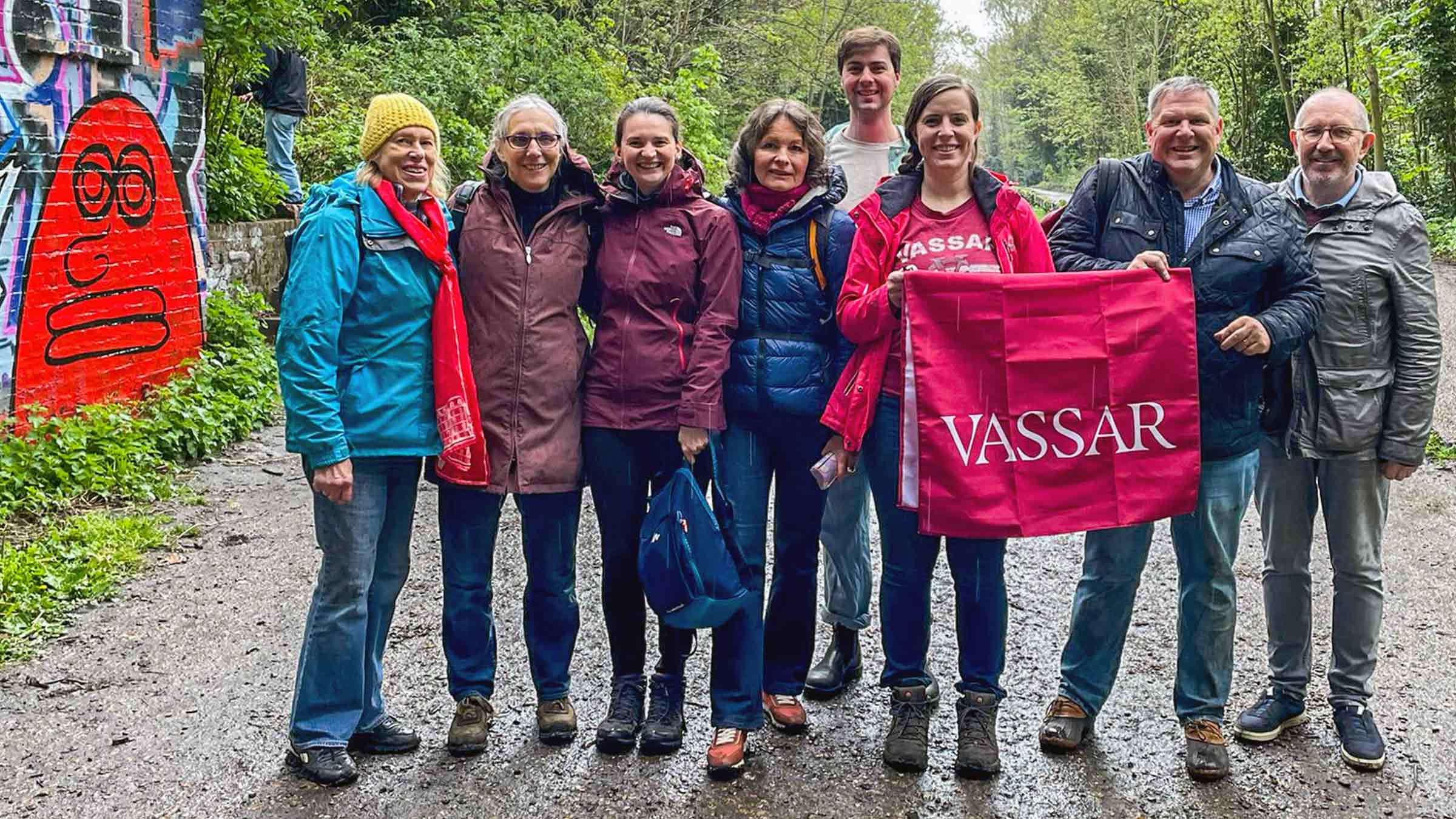 A group of people standing together on a gravel road in a wooded area holding a Vassar banner