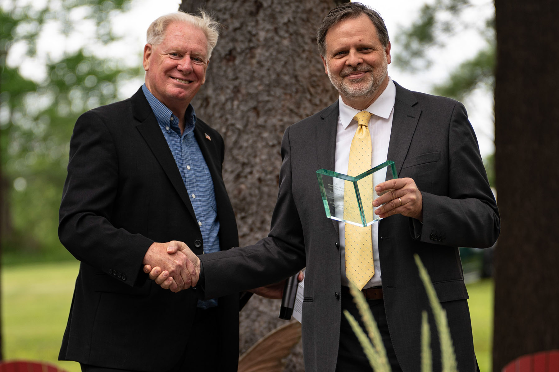 Two men standing in front of a tree shaking hands, one holding a clear glass award shaped like a book