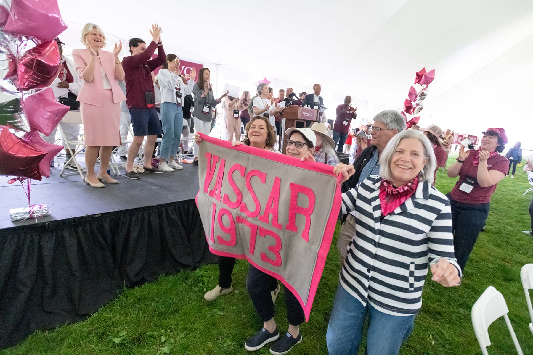 People standing on a small black stage on the lawn with other people walking by it holding a pink and gray flag saying "Vassar 1973"