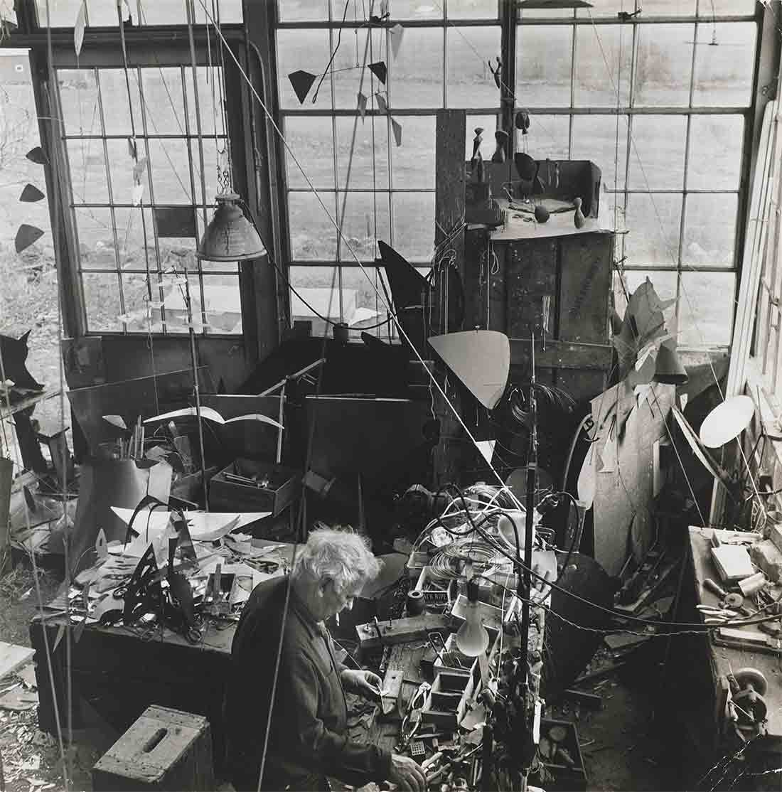 A person with unruly white hair stands in an extremely cluttered room with large windows, smoking a cigarette while working with some parts and pieces on a table completely filled with various objects.