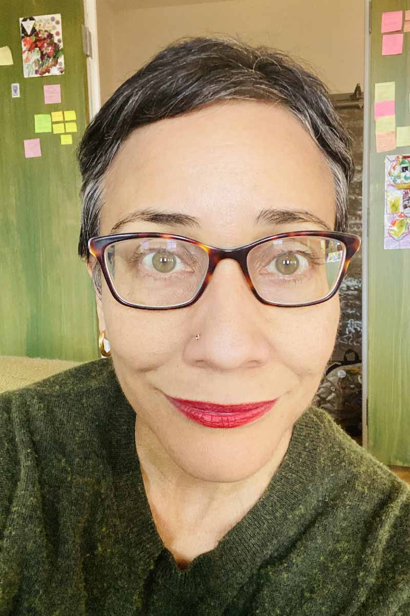 A close-up photo of a person with short dark hair, brown-rimmed glasses, and red lips smiling at the camera