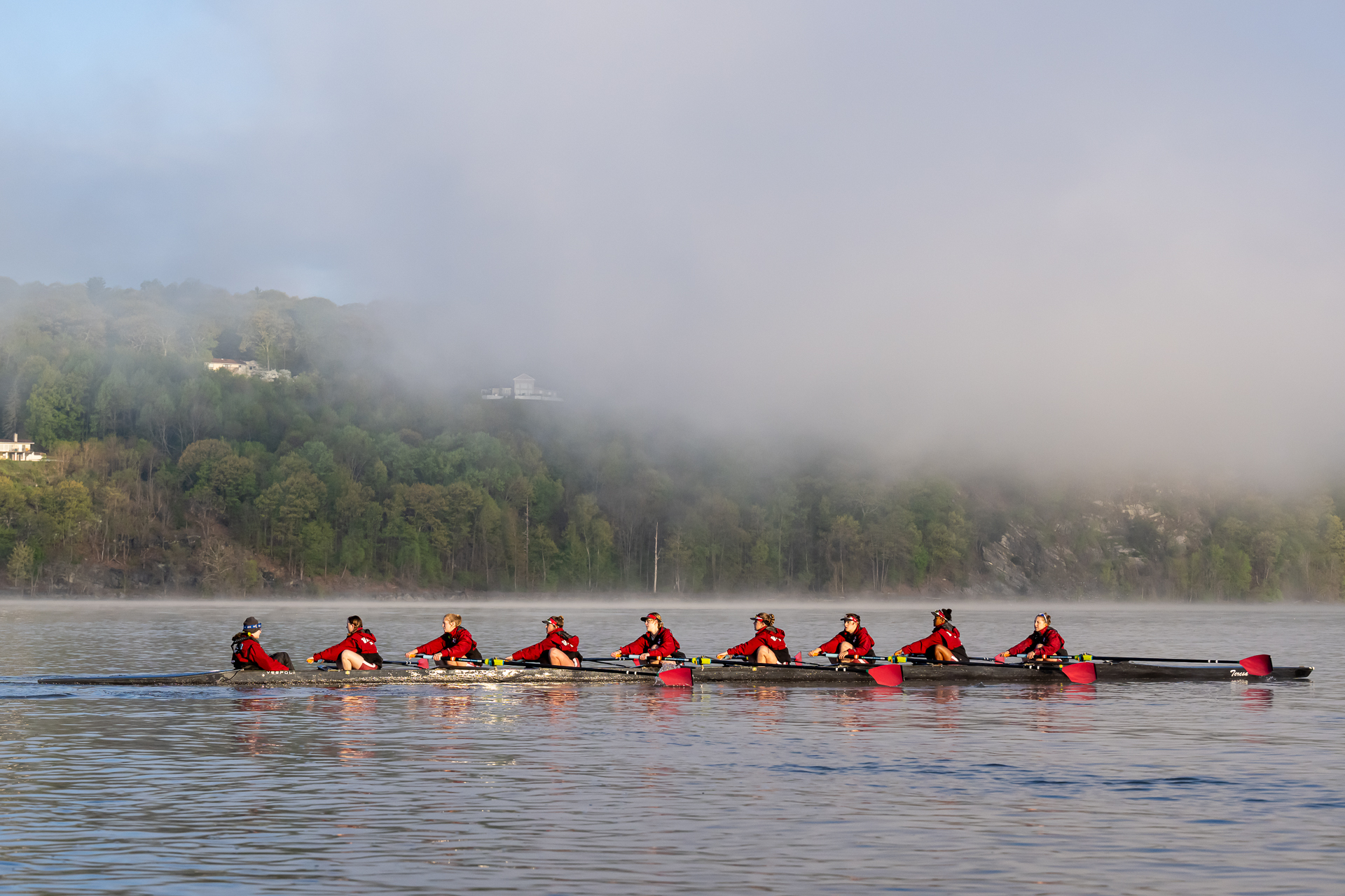 An 8-person boat rows on a river shrouded in morning mist.