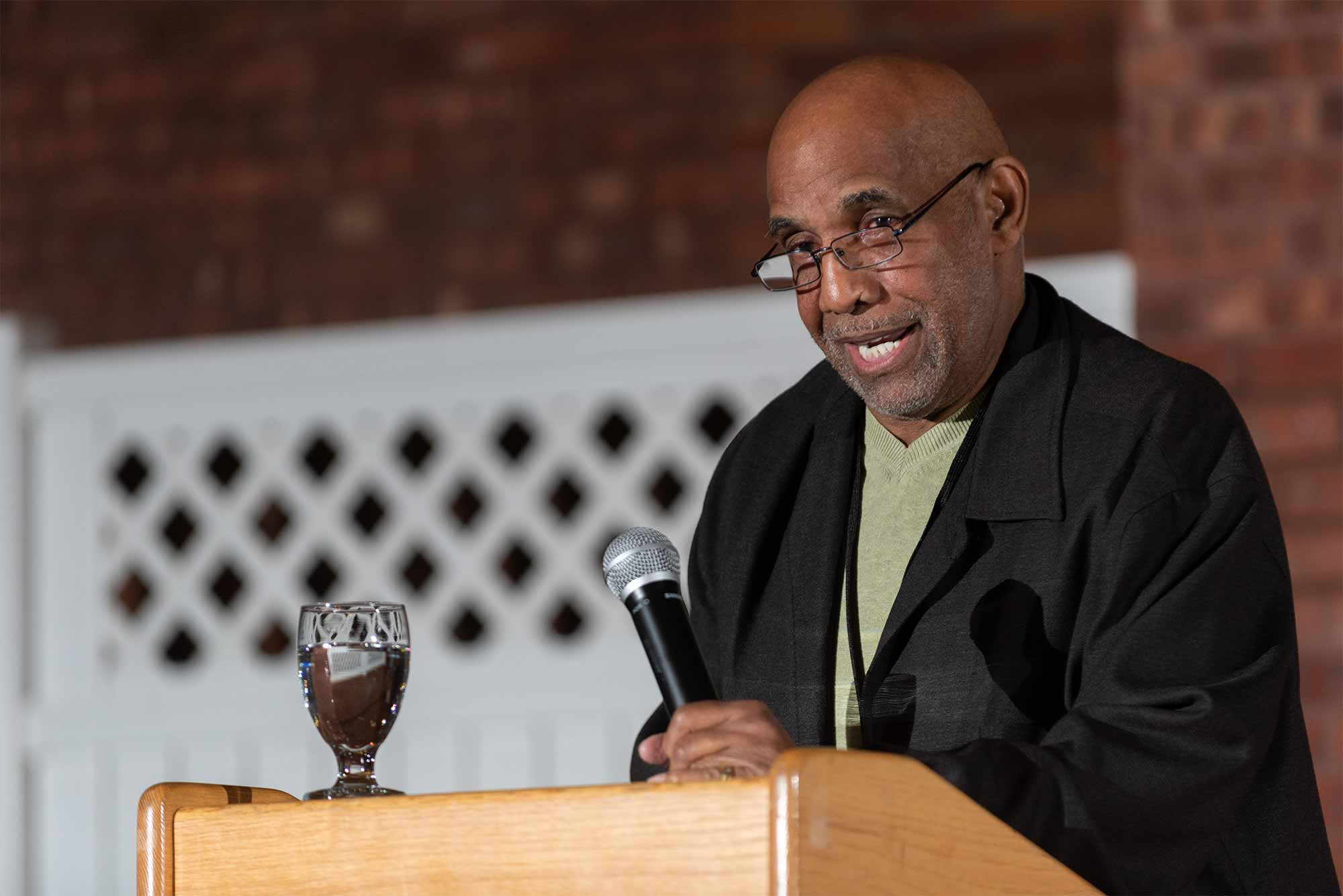 A person with a shaved head, light facial hair, and glasses speaks at a wooden podium, a brick wall visible in the background. The person is wearing a black formal jacket and a light-colored sweater.