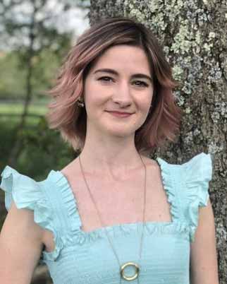 A person with long brown hair and a blue dress stands next to a tree, smiling at the camera.