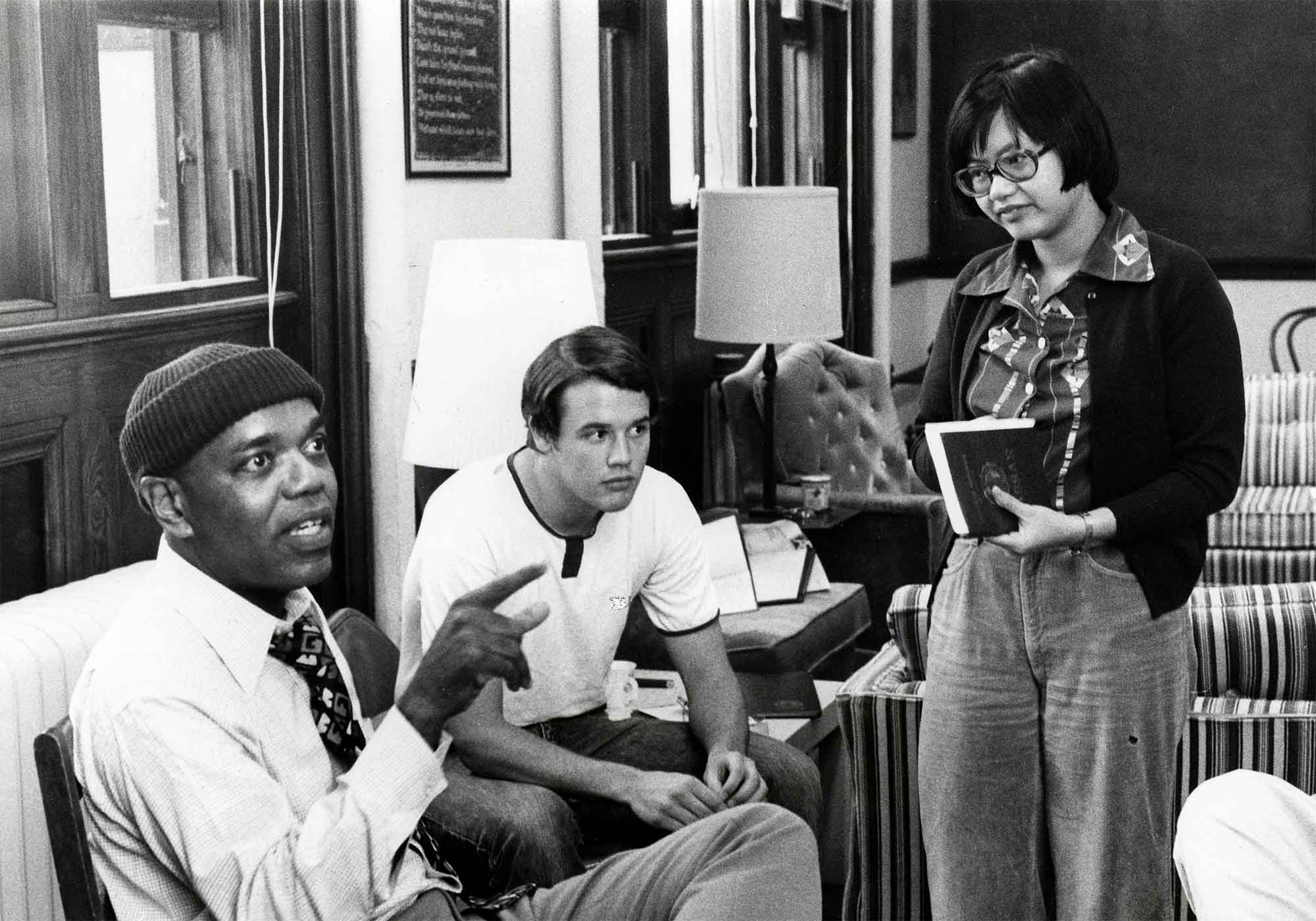 A black and white photograph three people in a room with furniture and lamps visible in the background. The person on the left is wearing a knit cap, a white shirt with a collar, and a patterned tie. The person is speaking with finger up. The middle person has short dark hair, a white shirt, and is looking away. The rightmost person is standing, has shoulder-length dark hair and glasses, and is looking at the speaker and holding a book.