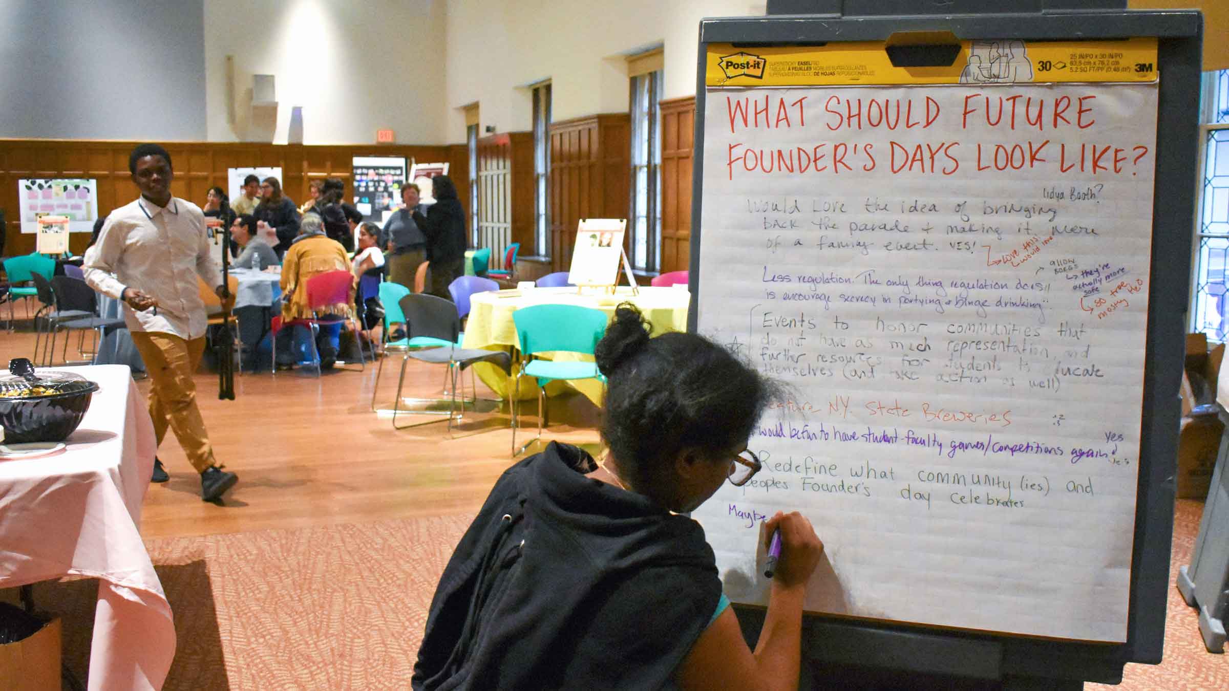 A very large room with wood-paneled walls and windows. An event is taking place here. In the foreground, a person with glasses and black hair in two buns is writing on a whiteboard, they’re back to the viewer. The whiteboard has a handwritten title at the top reading “What should future founder’s days look like?” Below it are written suggestions in different handwriting. In the background, a group of people sit at a table.