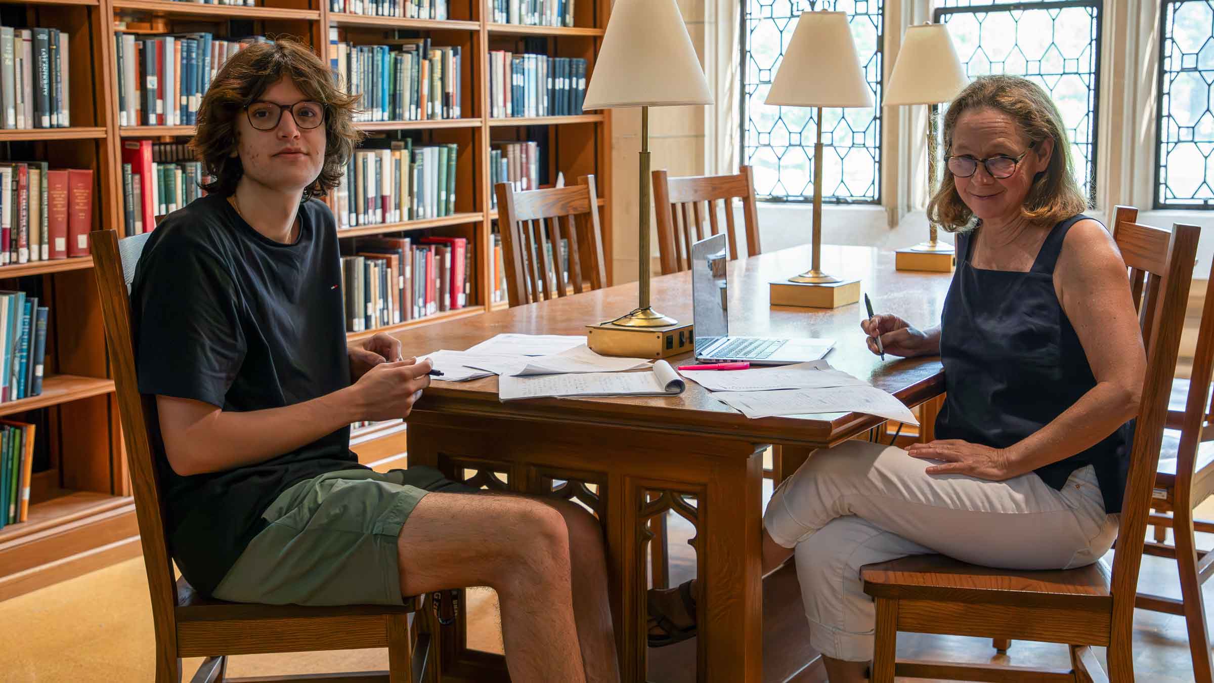 Two people seated at a desk with notepads and a laptop in a library