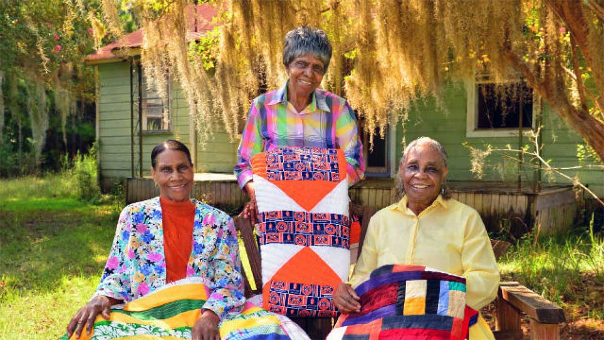 Three people, one standing and two sitting, in front of a house holding quilt work and smiling.