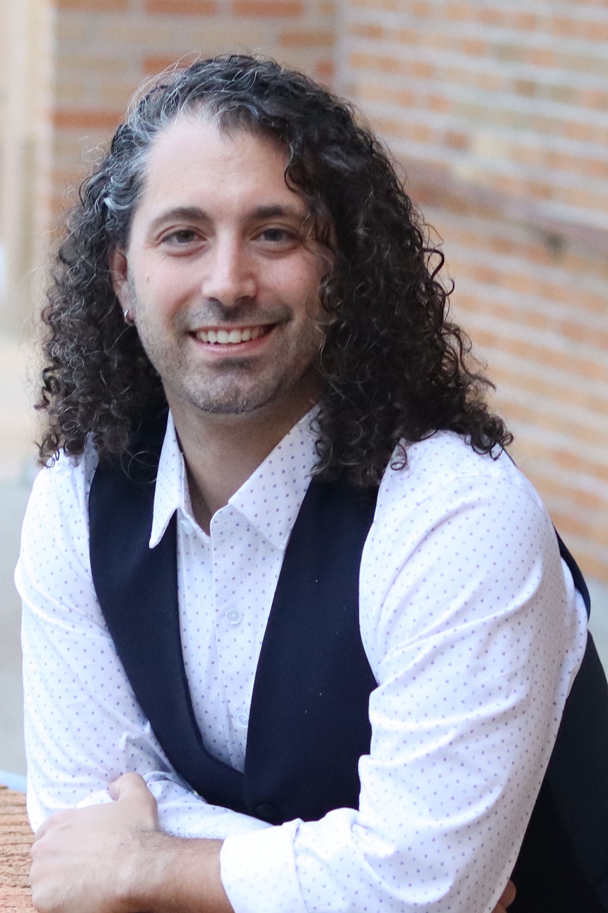 Person wearing white collared shirt and black vest with arms on a brick wall.