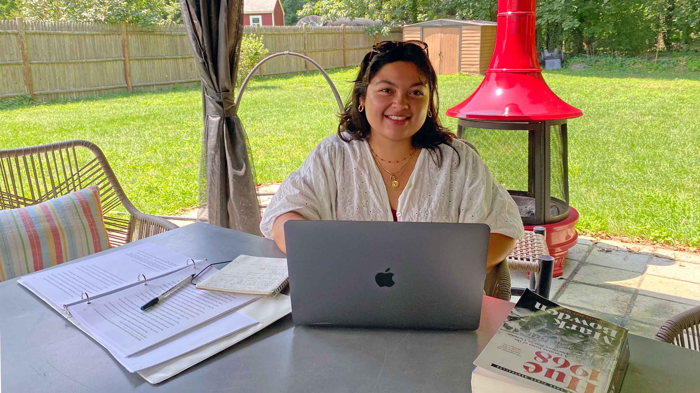 Person sitting at a table on a porch in the shade with a laptop, writing pad and a book smiling