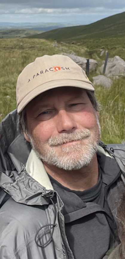A photo of a person smiling at the camera. The person has a gray beard and mustache, a tan baseball cap, and appears to be hiking on a mountain.