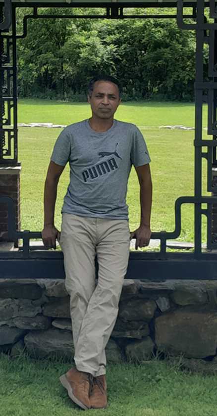 A person leans against a stone wall with a metal gate on it. The person has short black hair, a gray T-shirt with the word "Puma" on it, and tan slacks.