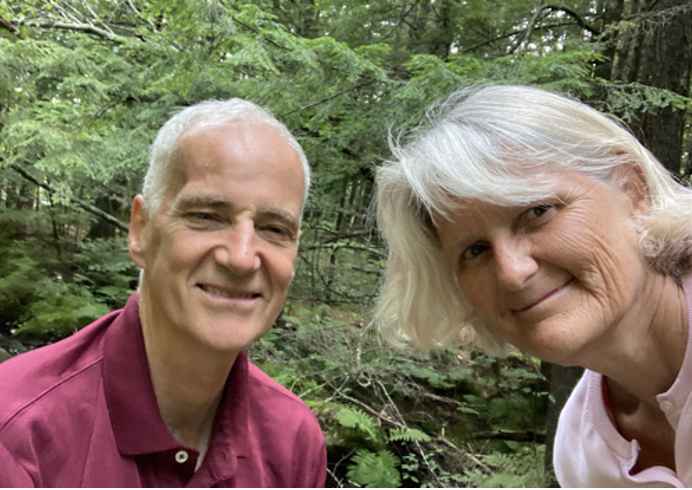Two people smile at the camera. The leftmost person has short gray hair and a maroon shirt. The rightmost person has shoulder-length gray hair and a pink shirt.