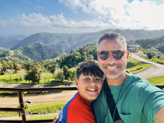 Two people smile at the camera. The leftmost person has glasses, short dark brown hair, and a red shirt. The rightmost person is taller with short gray hair, a gray beard and mustache, sunglasses, and a green shirt. Behind them is a mountain in the distance and a small rural road.