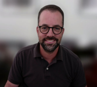 A person smiles at the camera. The person has short dark hair, glasses, and a dark beard and mustache. The background behind the person is blurred, suggesting they are on a Zoom call.