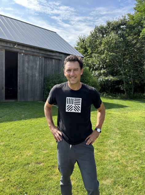 A person stands outside on a grassy area, with trees and a barn in the background. The person has short dark hair, a black T-shirt with an abstract geometrical design on it, and gray slacks.