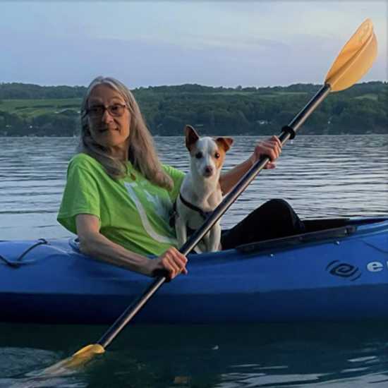 A person kayaking in a river. The person has long gray hair, glasses, and a bright green T-shirt. Sitting in the front of the kayak on the person's lap is a small brown and white dog.