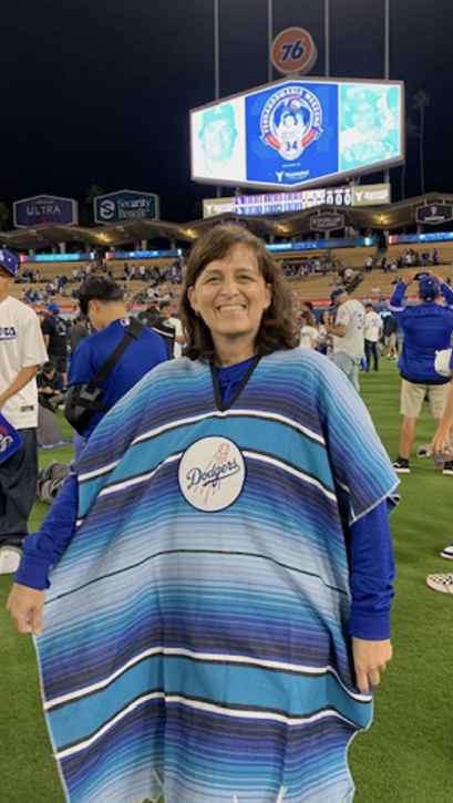 A person standing in the middle of a sports field at night, with fans and baseball players in the background. The person is wearing a long, blue-striped dress with "Dodgers" on it. Behind them is a large illuminated stadium sign. It's not clear what's on the sign.