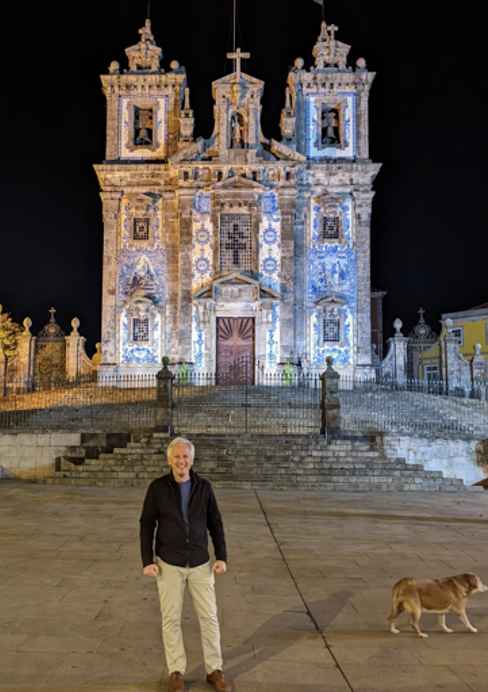A person standing in front of an ornate, smallish cathedral at night. The person has short gray hair, a black shirt, and tan slacks. A golden retriever is walking near them.