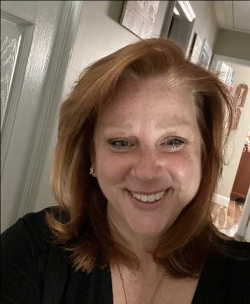 A person with long, auburn hair and a black shirt. The person is smiling at the camera, and is standing in the hallway of a house.