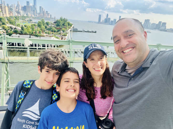 Four people smile at the camera. The leftmost person has short brown hair and a dark gray T-shirt with the words "North Cove Sail KIDSail" partially visible. The second person has short brown hair and a blue shirt. The third person has long brown hair, a pink shirt, and a blue baseball cap with "SLS" on it. The rightmost person is taller than the others, has very short dark hair, and a gray shirt. A city and harbor are visible in the background.