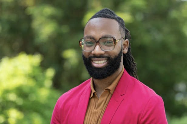 A person smiles at the camera. The person has long black, curly, braided hair, a black mustache, glasses, and a bright pink shirt. The person is standing outdoors; blurry foliage is visible in the background.