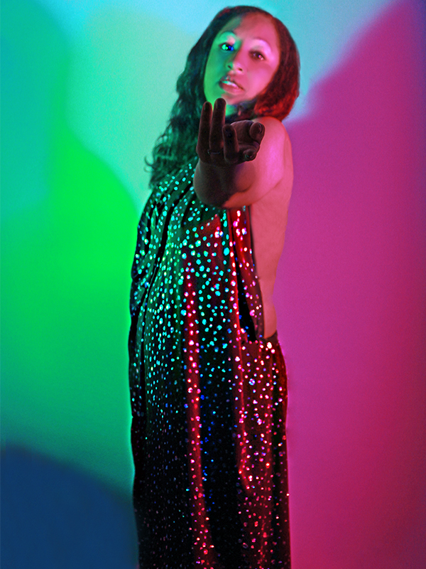 A women in a sparkly gown stares at the camera while holding her hand out in a beckoning pose.