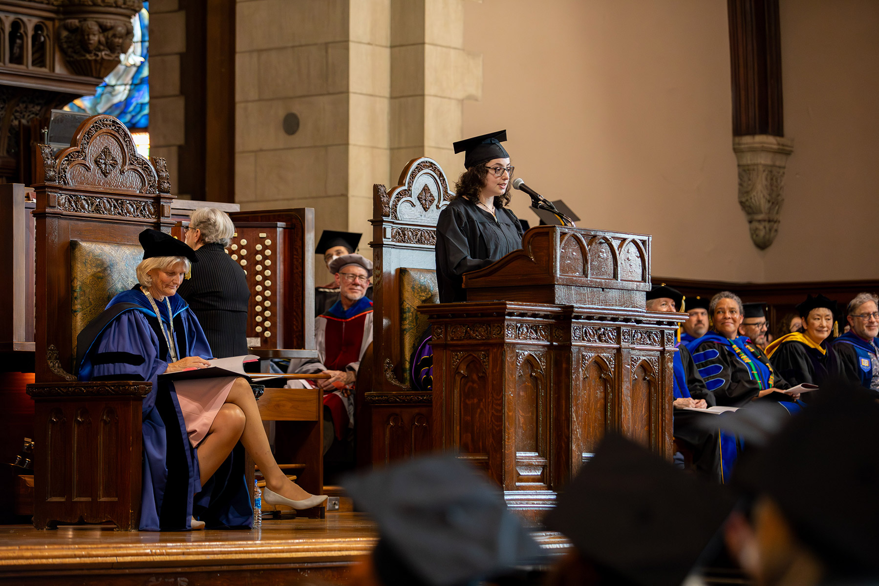 Person standing at a podium with a woman sitting in a large wooden throne behind her with professors in the background looking on.