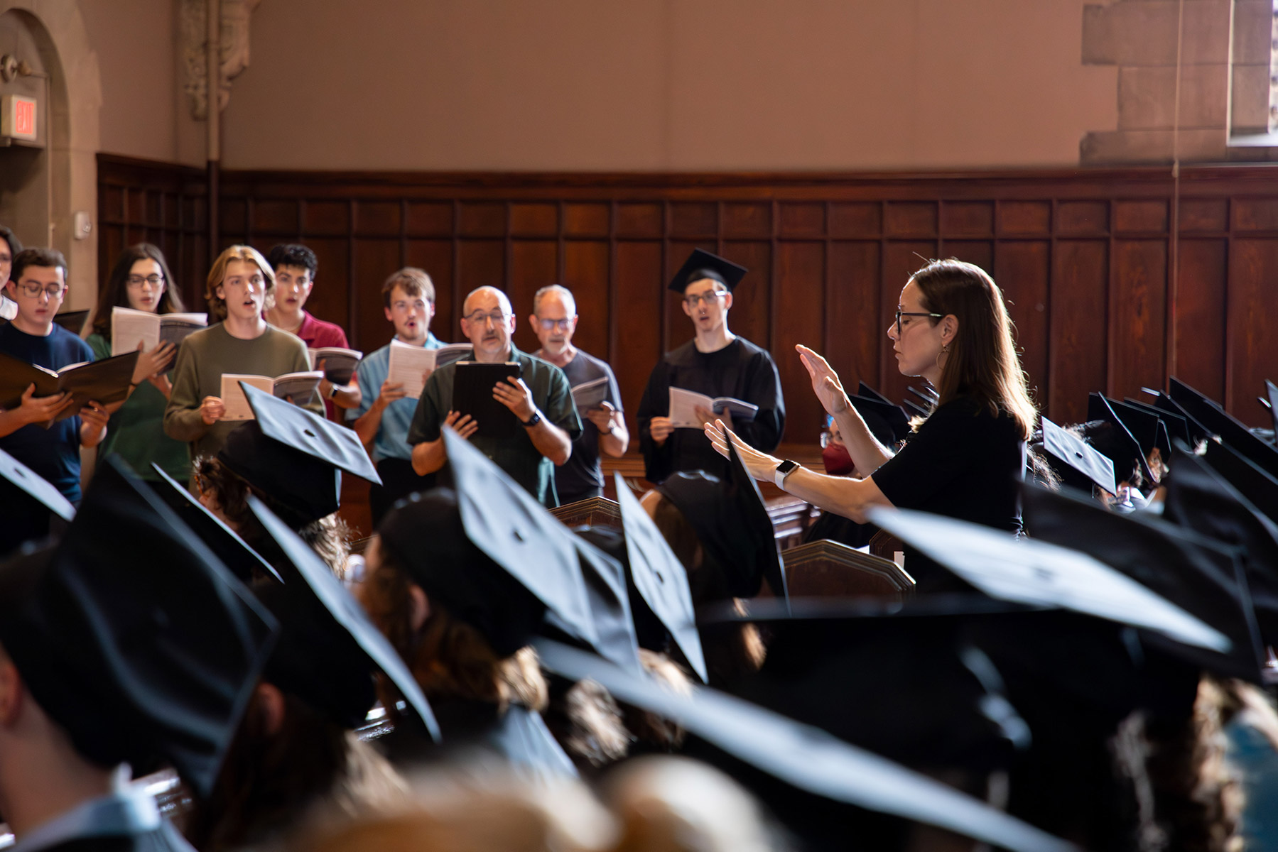 Conductor standing gesturing with singers in the background and students with graduation caps in the foreground.