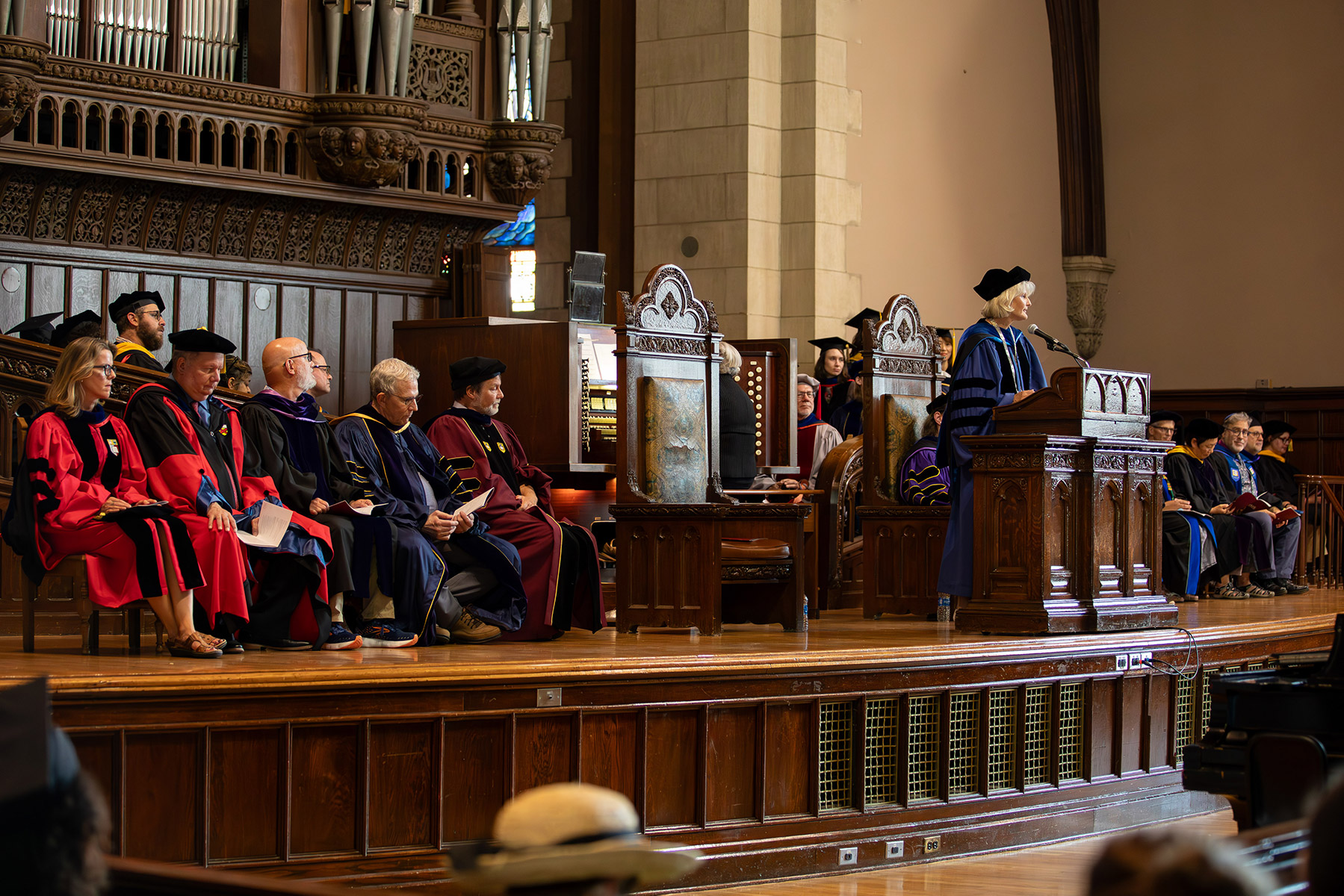 Person standing at a podium with professors sitting behind them and students in graduation caps sitting in front.