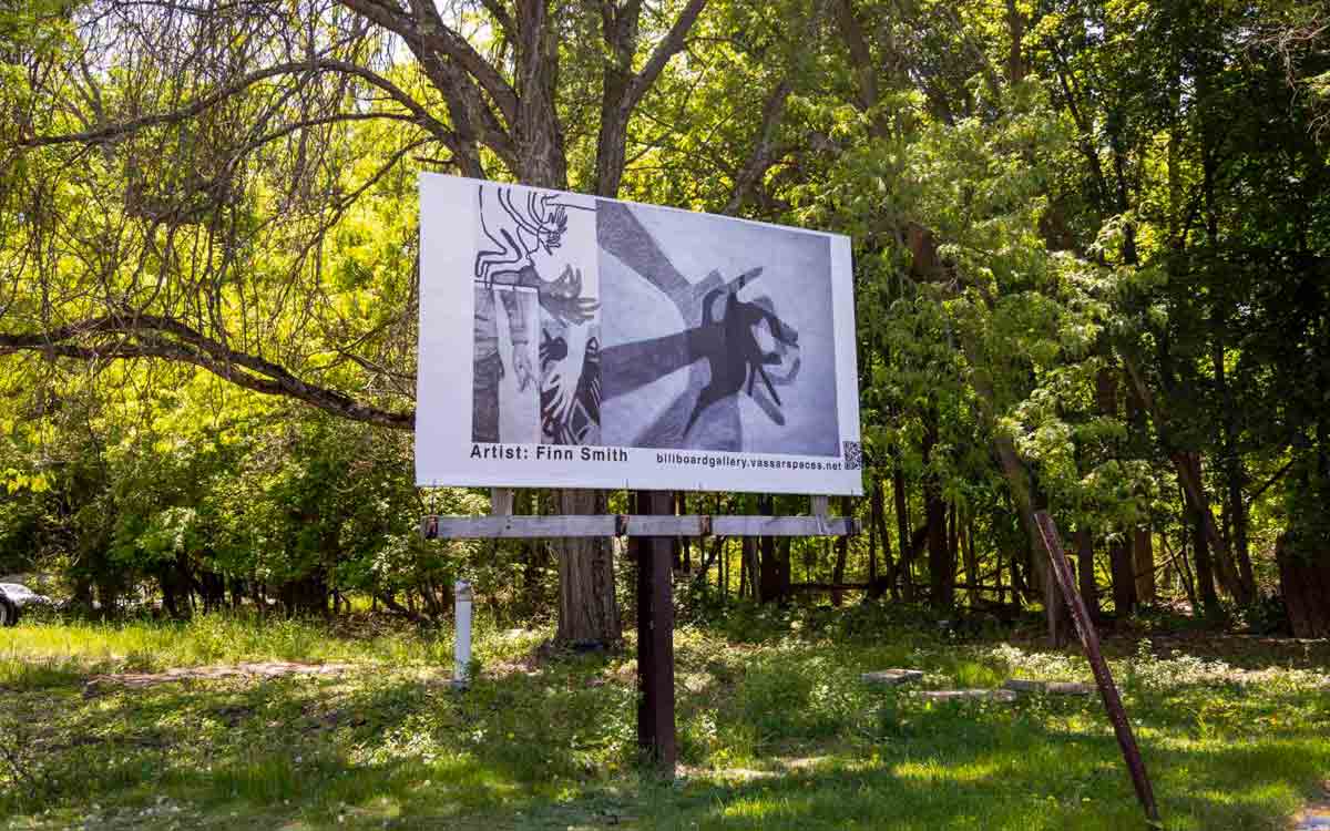 A billboard stands in a wooded, sunlit area. The billboard displays several monochrome abstract images of hands. Below is the text “Artist: Finn Smith”, followed by “billboardgallery.vassarspaces.net”.