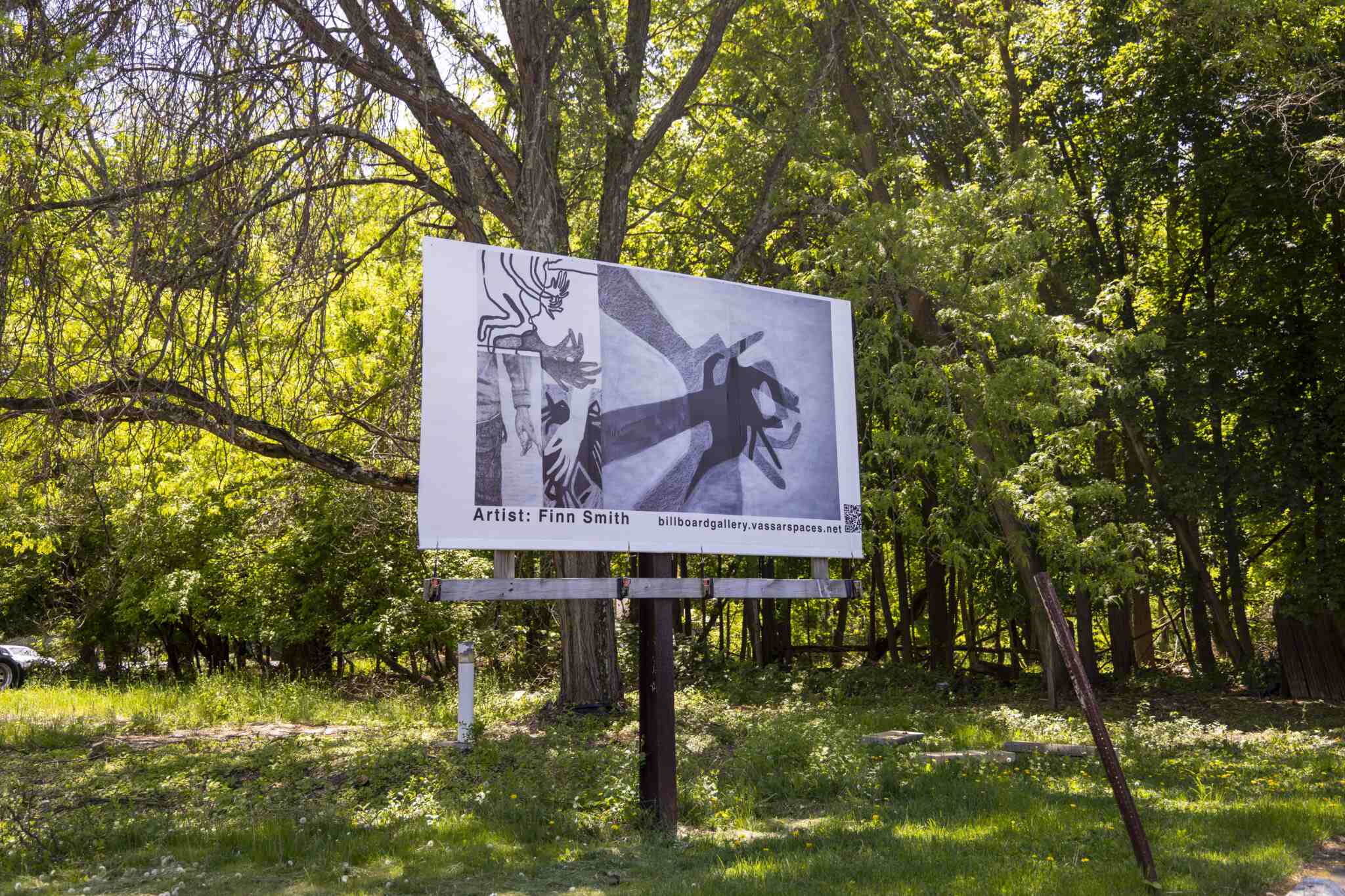 A billboard stands in a wooded, sunlit area. The billboard displays several monochrome abstract images of hands. Below is the text “Artist: Finn Smith”, followed by “billboardgallery.vassarspaces.net”.