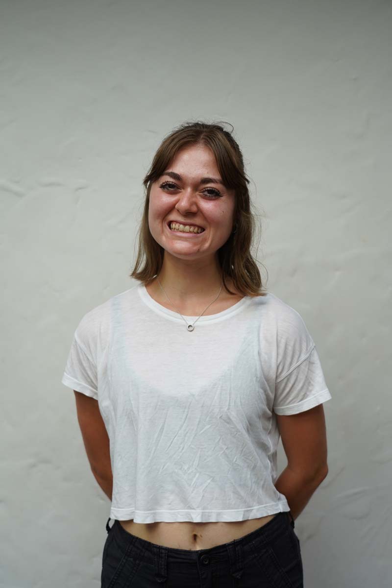 A person with long dark blond hair and a white T shirt smiles at the viewer.