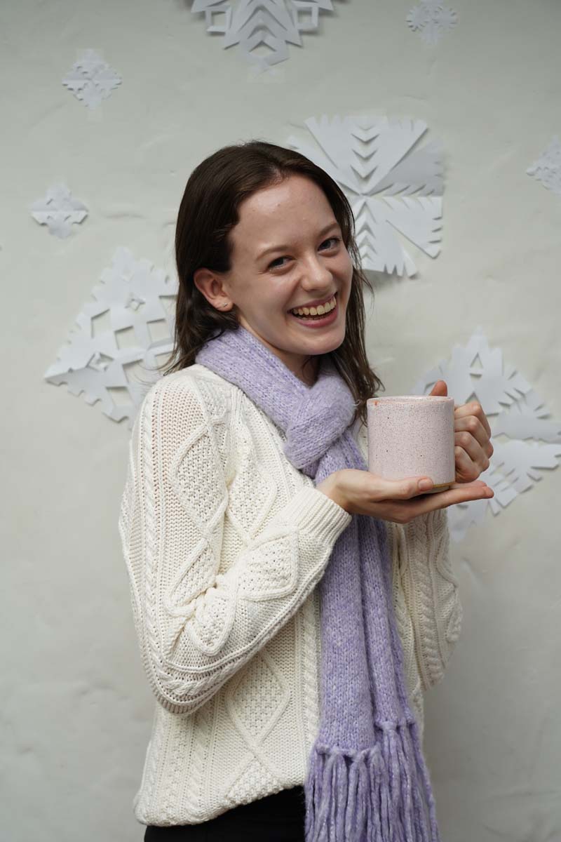 A person with long dark brown hair and a white winter coat with a purple scarf smiles at the viewer while standing in front of a wall of paper snowflakes.