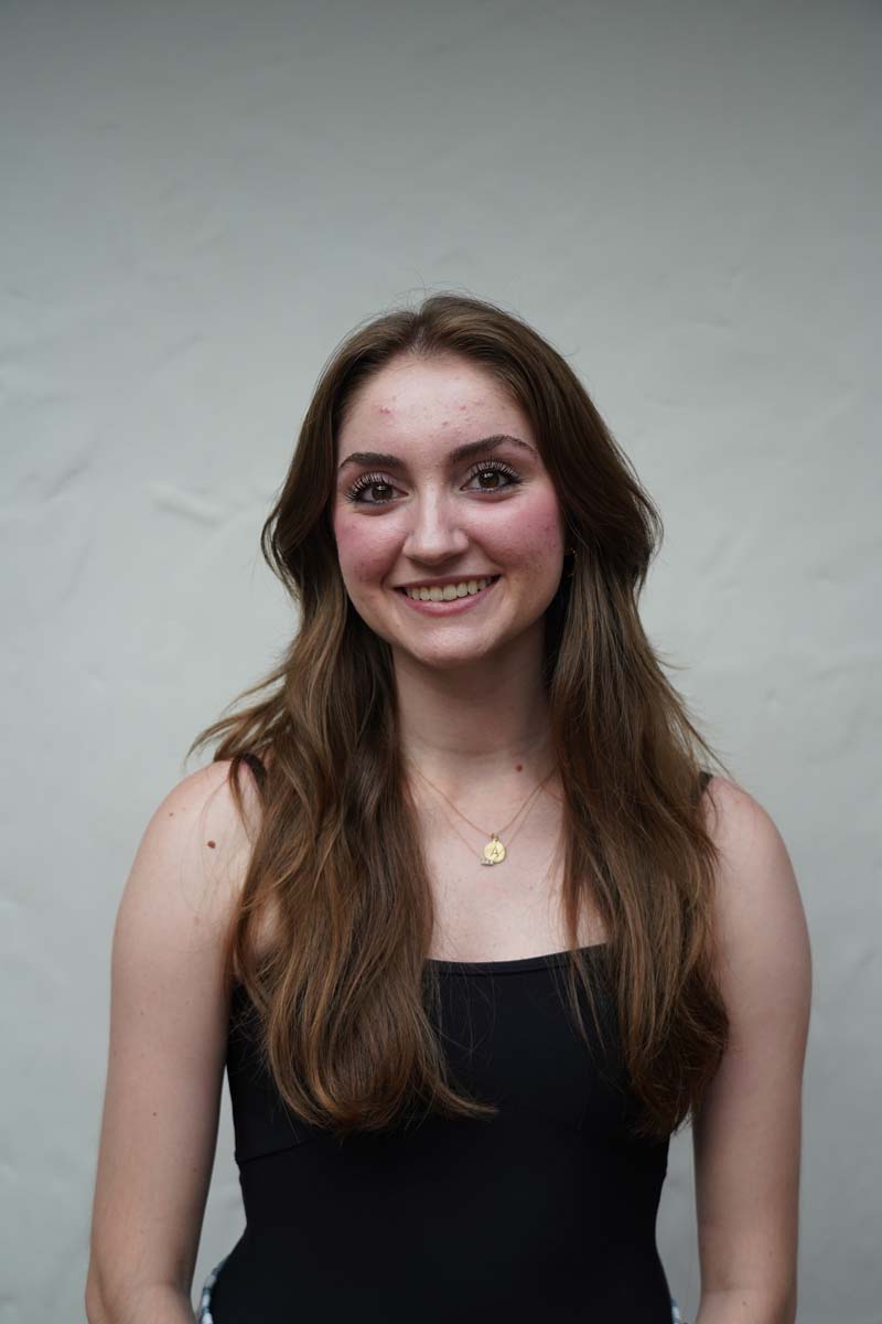 A person with long auburn hair and a black tank top smiles at the viewer.